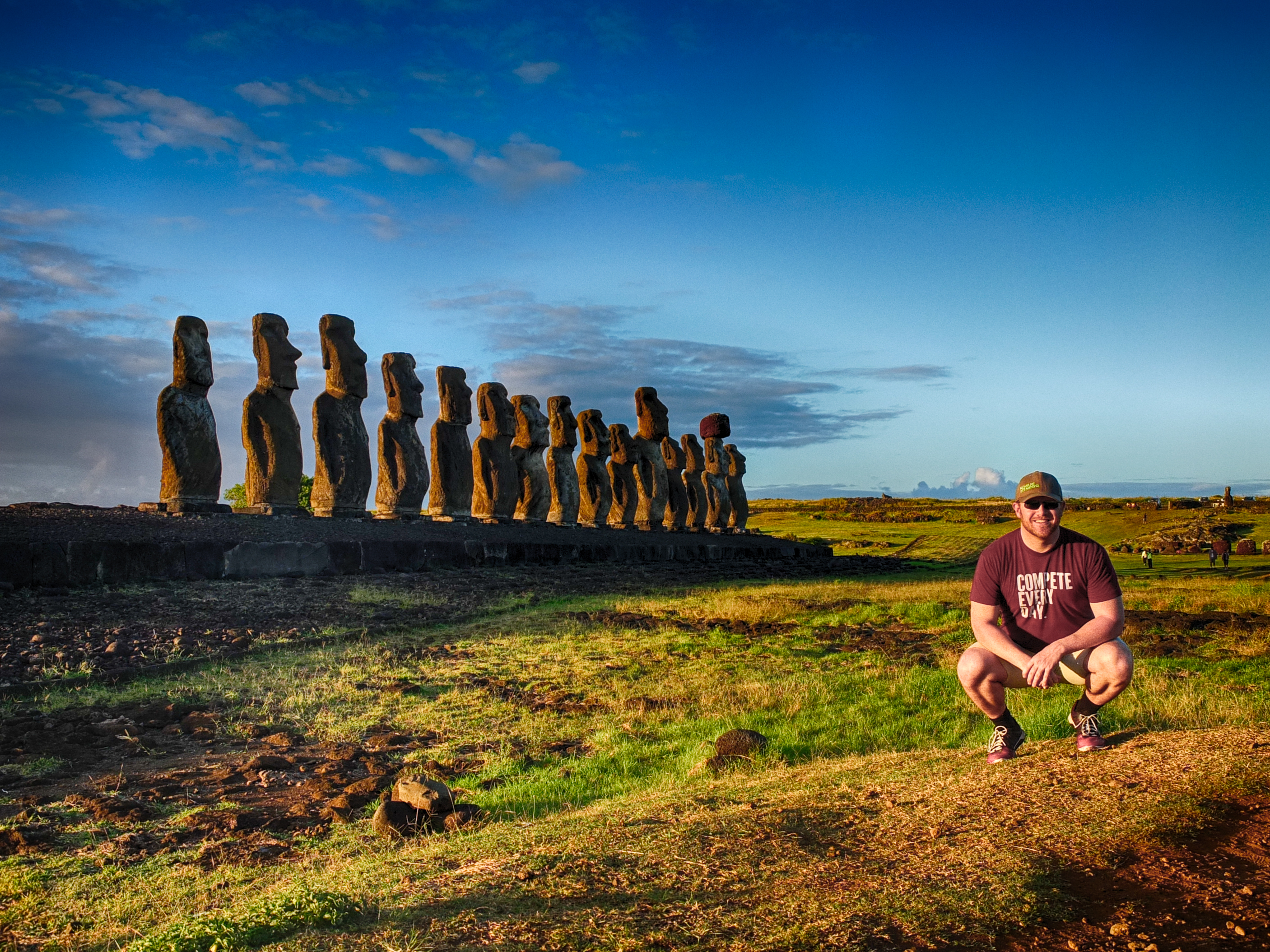a man squatting in front of a row of statues with Easter Island in the background