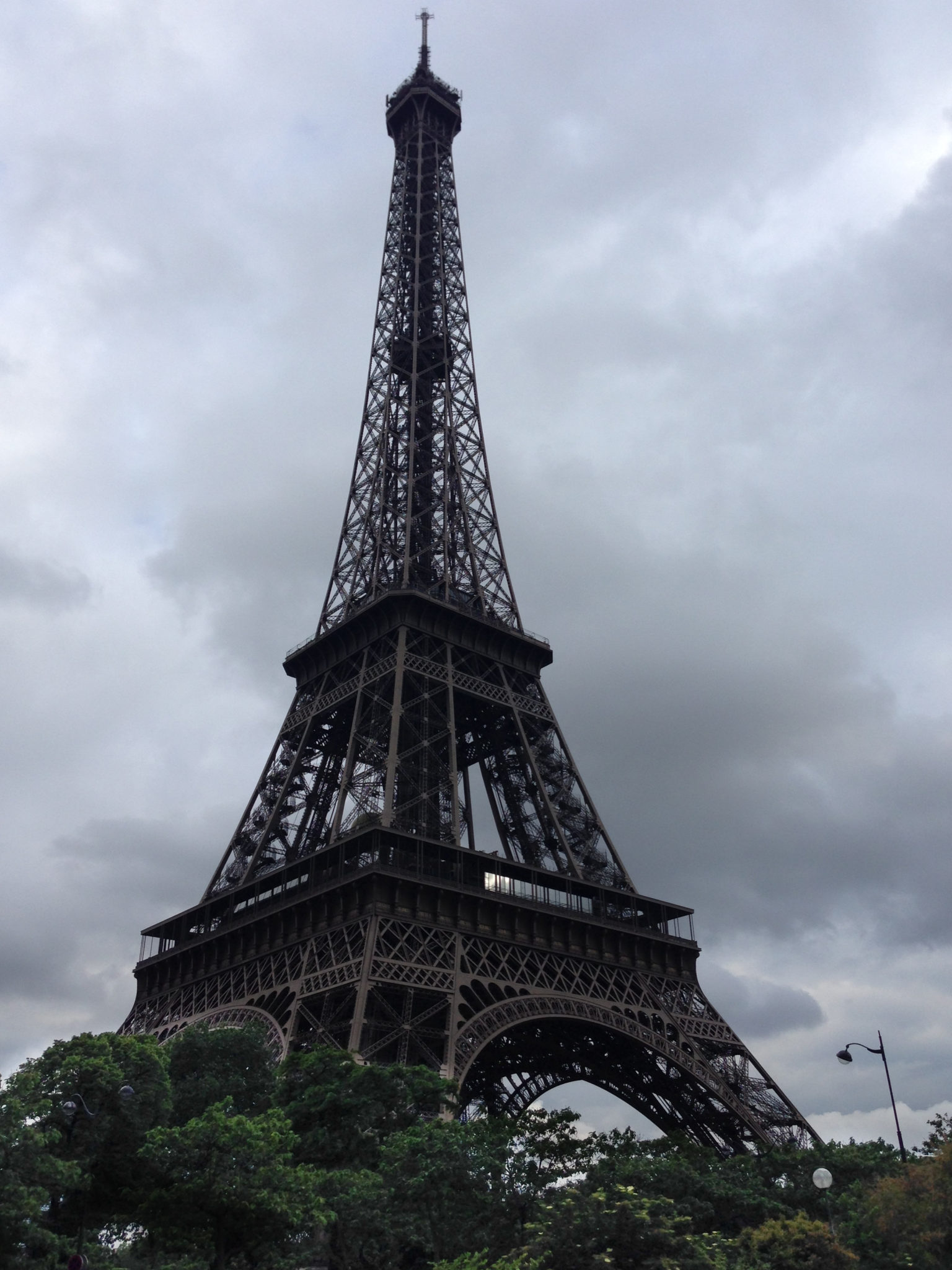 a tall metal tower with trees and a cloudy sky with Eiffel Tower in the background