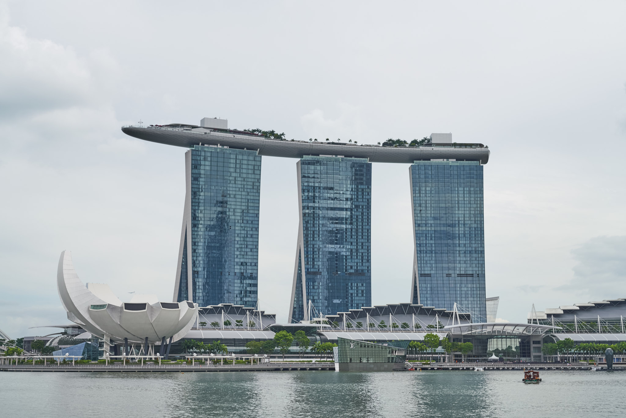a group of tall buildings with a boat floating above them with Marina Bay Sands in the background