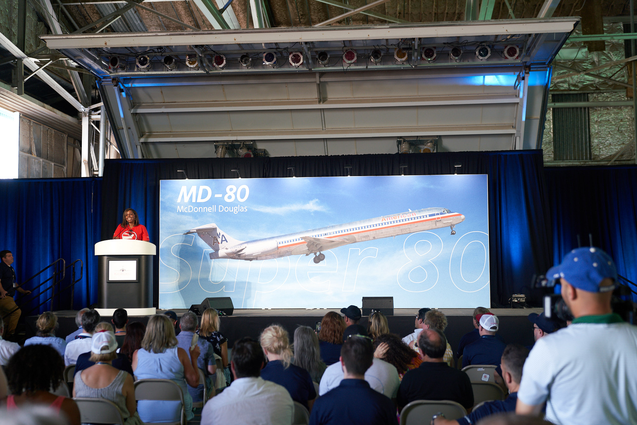 a woman standing at a podium in front of a large screen with a large airplane on it